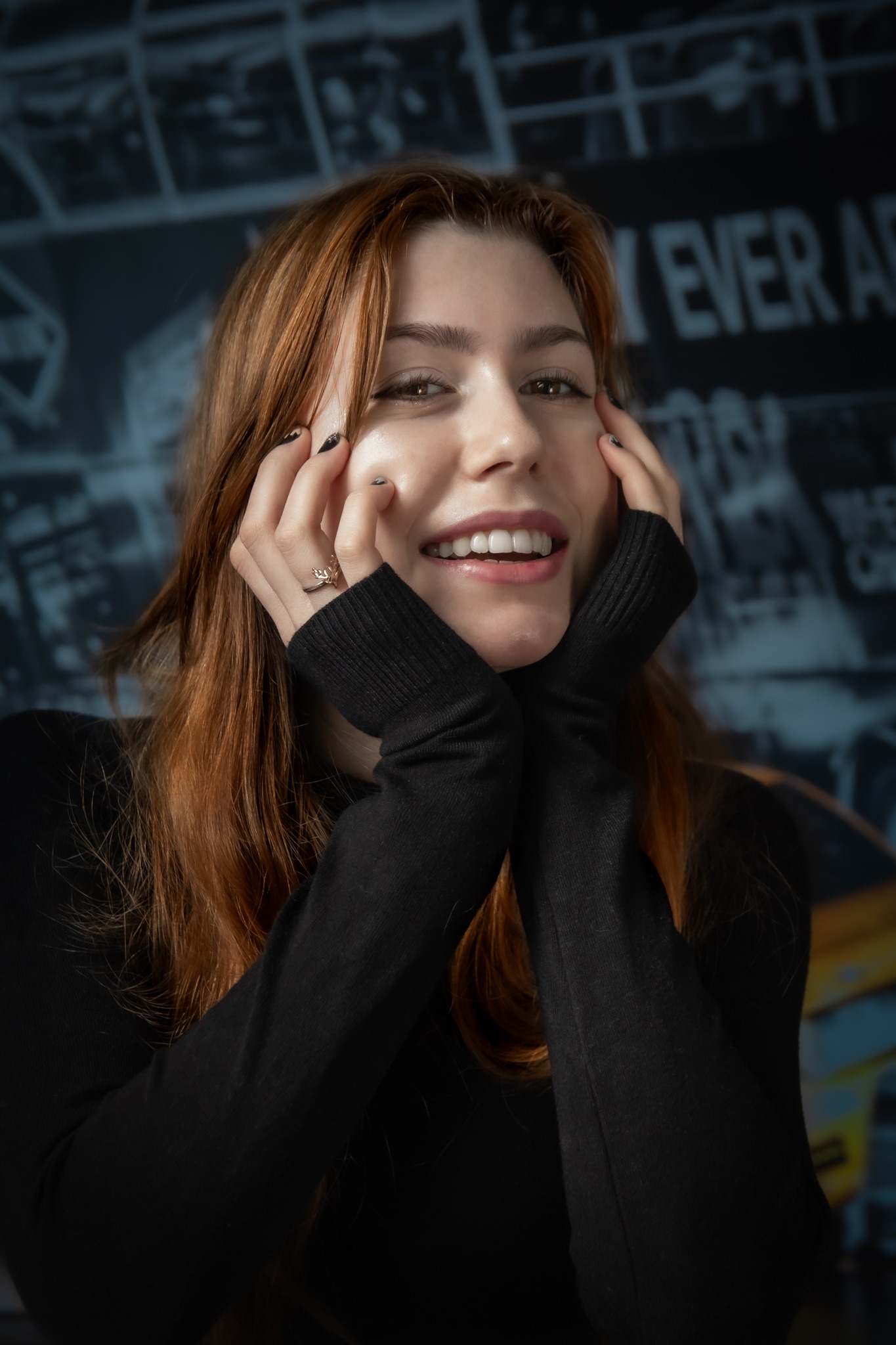 Portret uśmiechniętej rudowłosej kobiety z dłońmi przy twarzy, w czarnym golfie, na tle ściany z grafiką miejską w Gdańsku. Widoczny pierścionek na palcu.