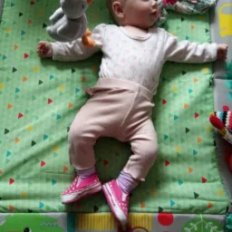 Niemowlę leży na zielonej macie edukacyjnej z kolorowymi trójkątami, ubrane w różowe spodnie i białą bluzkę w kropki, na stopach ma różowe buciki, a nad nim wisi pluszowa zabawka.