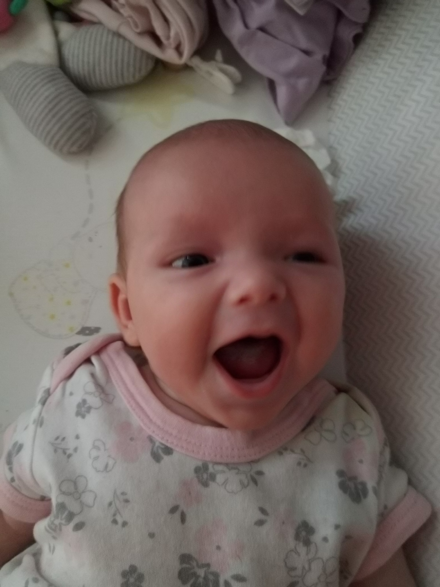 Uśmiechnięte niemowlę w białym ubranku z różowymi wykończeniami i kwiatowym wzorem leży na wzorzystej pościeli obok pluszowych zabawek.