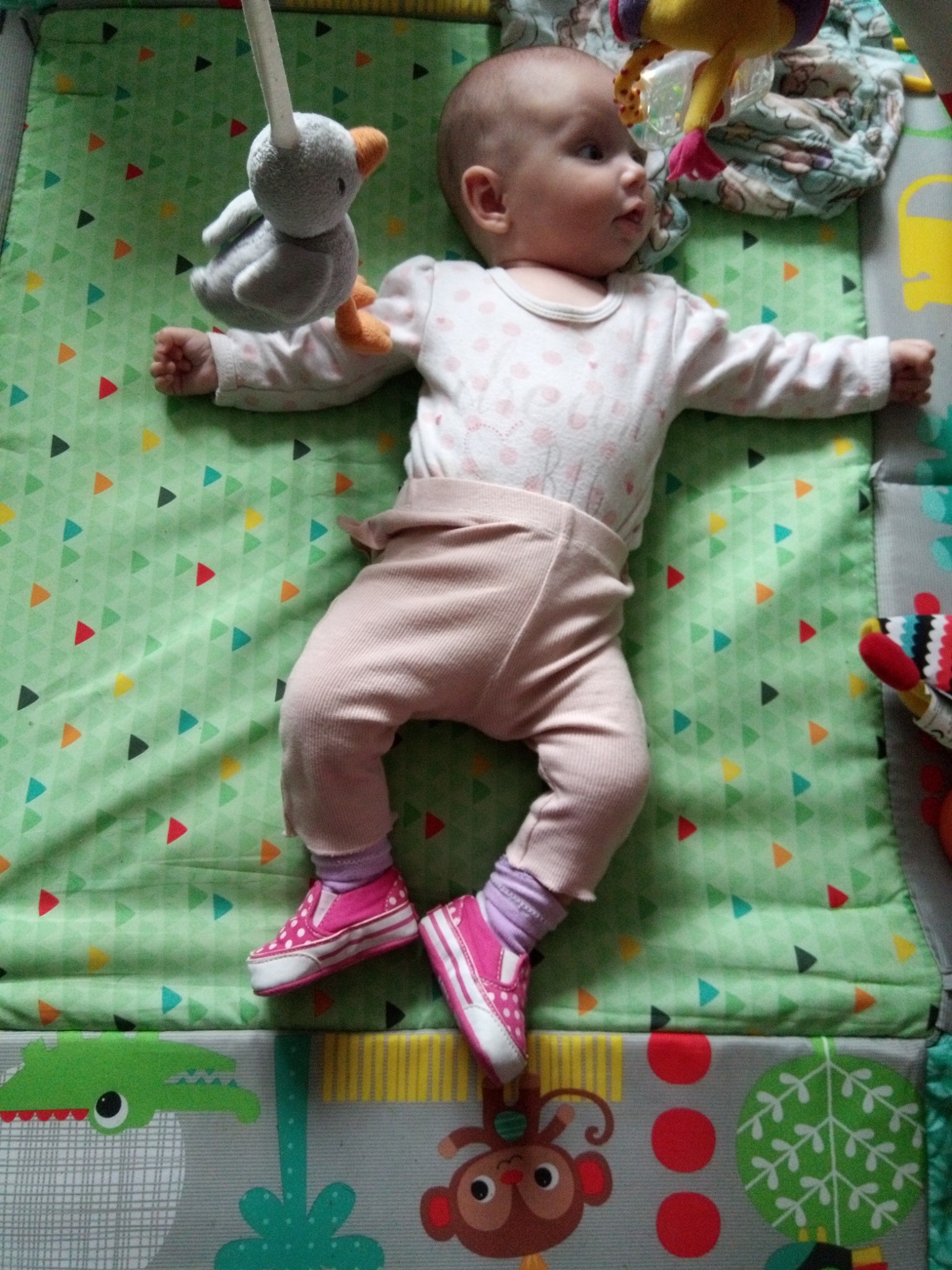 Niemowlę leży na zielonej macie edukacyjnej z kolorowymi trójkątami, ubrane w różowe spodnie i białą bluzkę w kropki, na stopach ma różowe buciki, a nad nim wisi pluszowa zabawka.