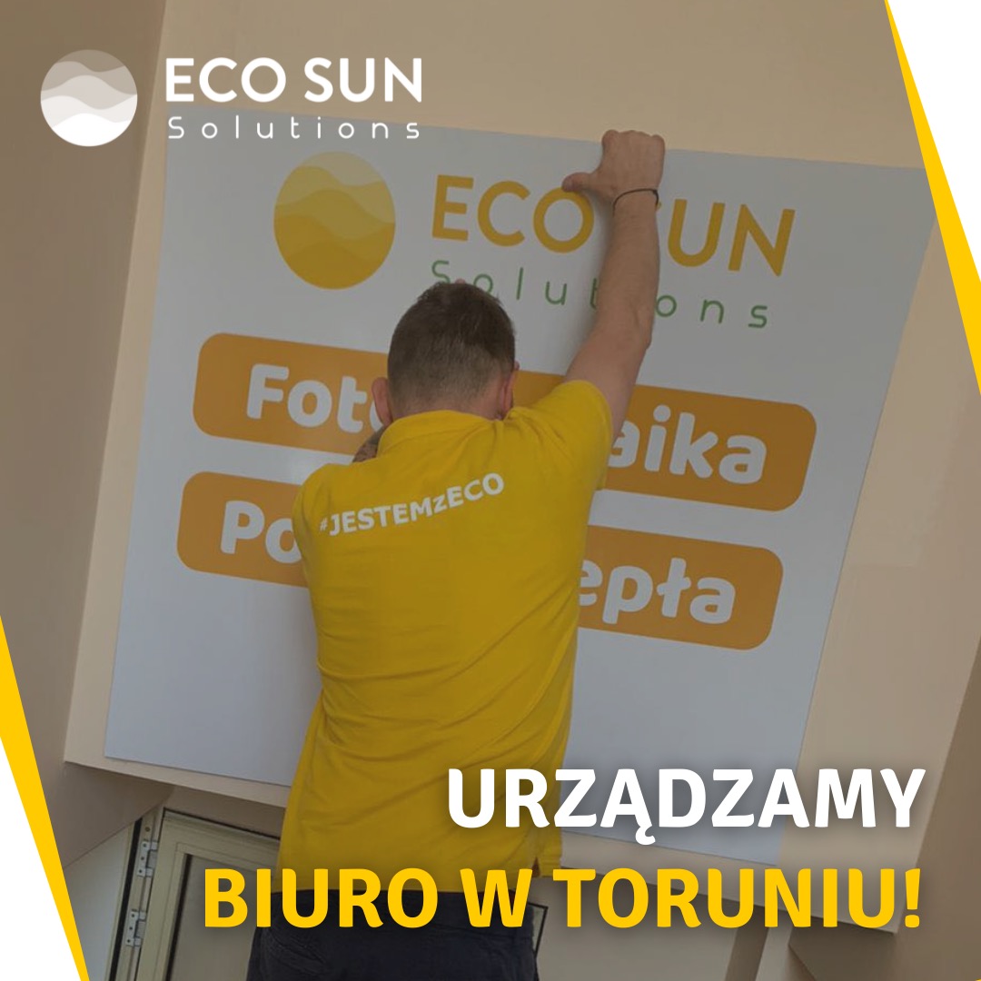 Mężczyzna w żółtej koszulce z napisem 'JESTEMZECO' montuje duży baner reklamowy z napisem 'ECO SUN Solutions', 'Fotowoltaika' i 'Pompy ciepła' w nowo urządzanym biurze w Toruniu.
