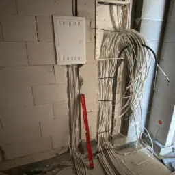 Wewnętrzne okablowanie elektryczne w surowym stanie, widoczna skrzynka rozdzielcza, kable prowadzone w peszlach i mocowane do ściany z betonu komórkowego, poziomica oparta o ścianę.