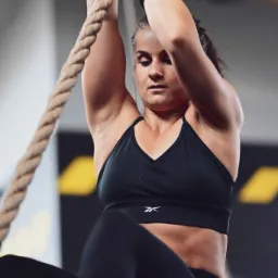 Kobieta w czarnym sportowym stroju wspina się po linie, używając rąk zabezpieczonych czarnymi opaskami, widoczny wysiłek na twarzy, tło szare z elementami żółci.