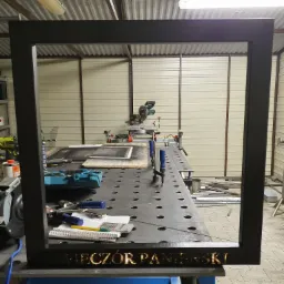 Czarna, metalowa rama z wyciętym napisem 'WIECZÓR PANIEŃSKI' stoi na stole warsztatowym, w tle narzędzia i piła do metalu.