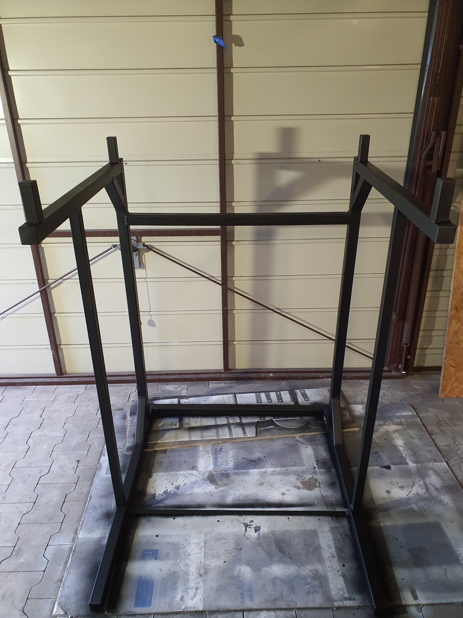 Stalowa konstrukcja ramy z profili zamkniętych, świeżo pomalowana na czarno, widoczna na tle zamkniętej bramy garażowej z poziomymi przetłoczeniami.