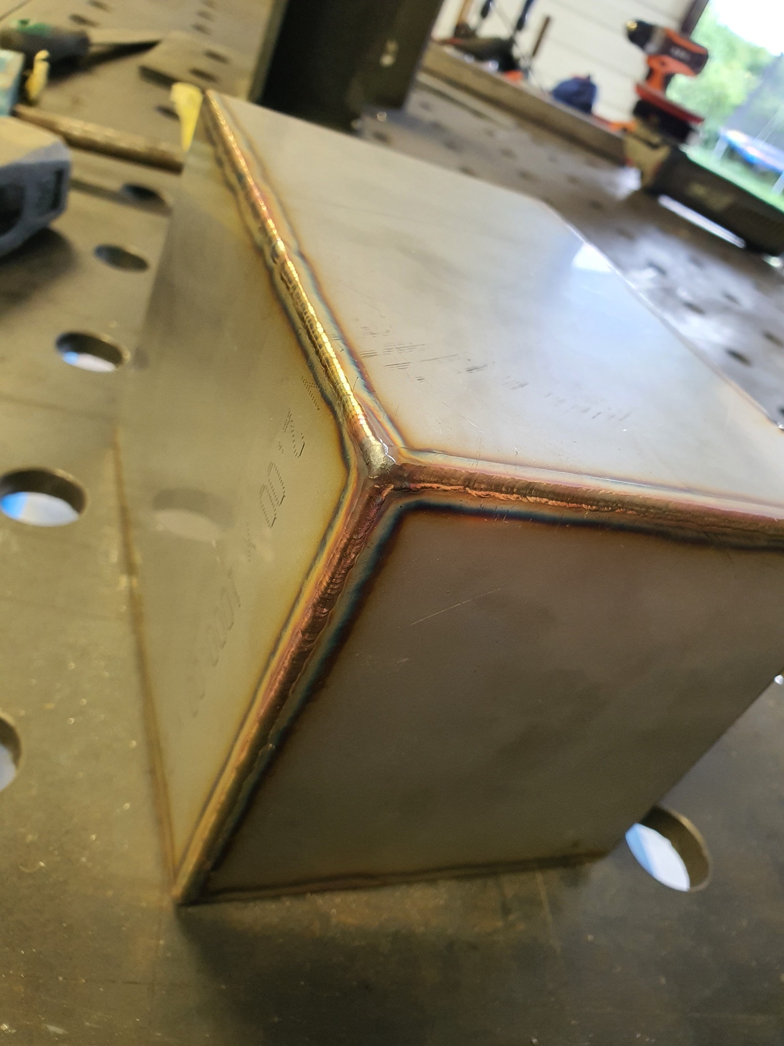 Metalowy prostopadłościan z widocznymi śladami spawania na krawędziach, leżący na perforowanym stole warsztatowym z narzędziami w tle.