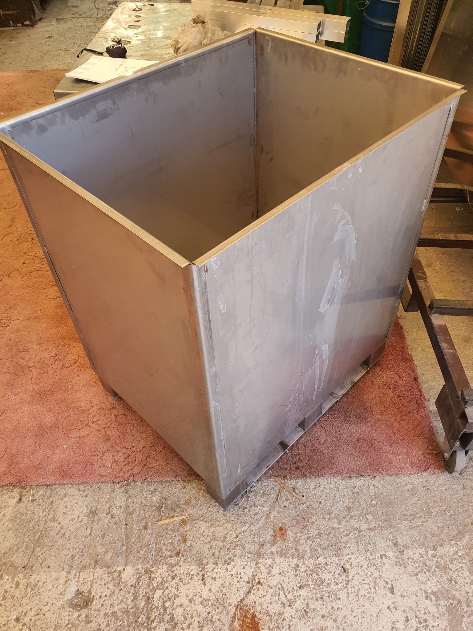 Duży, surowy, metalowy pojemnik w kształcie ściętego ostrosłupa, spawany z blachy, z widocznymi śladami obróbki, na podłodze warsztatu.