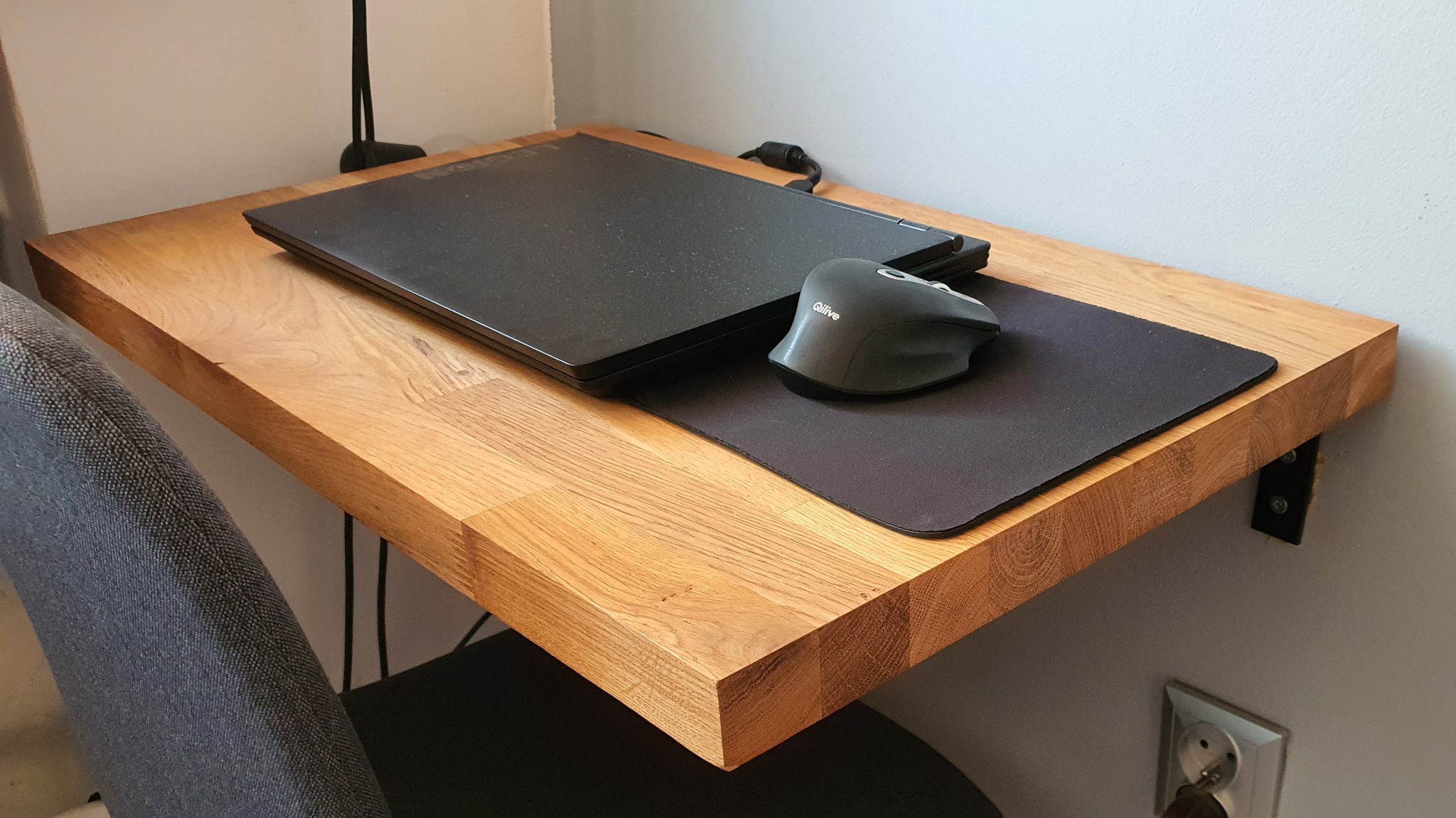 Przyścienny, dębowy blat biurka z widocznymi mocowaniami, na którym leży zamknięty laptop, myszka bezprzewodowa i podkładka pod mysz.