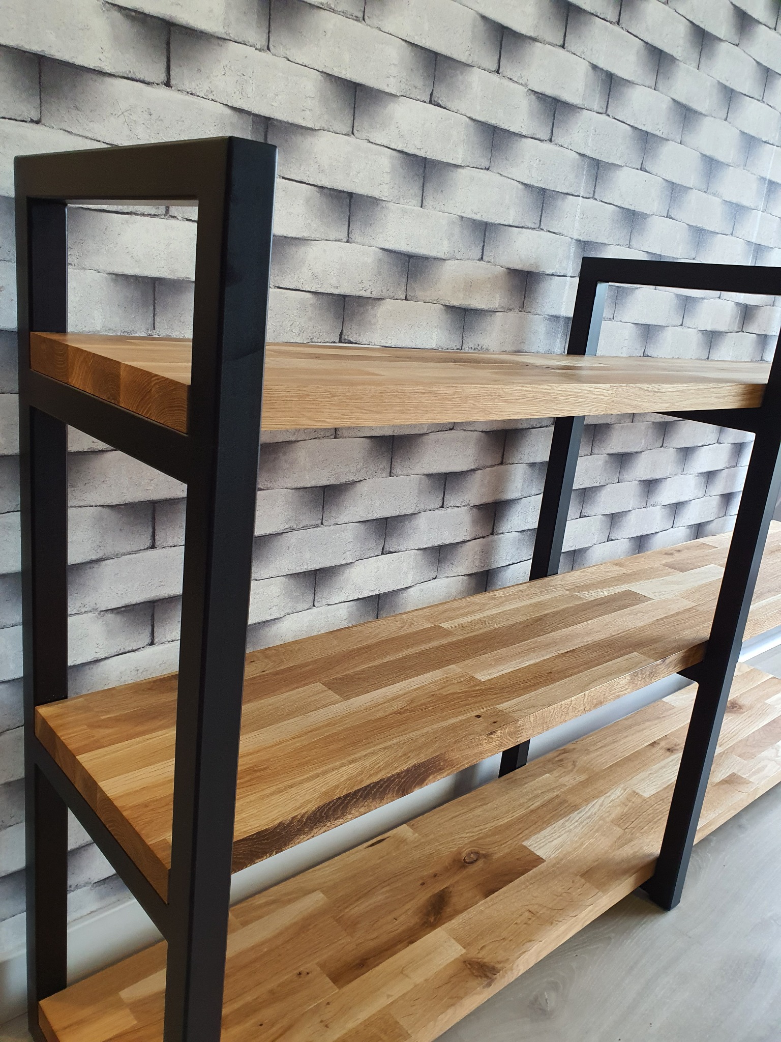 Ręcznie wykonany regał z drewnianymi półkami o jasnym odcieniu i czarną, metalową ramą, stojący na szarej podłodze na tle ściany imitującej cegłę.