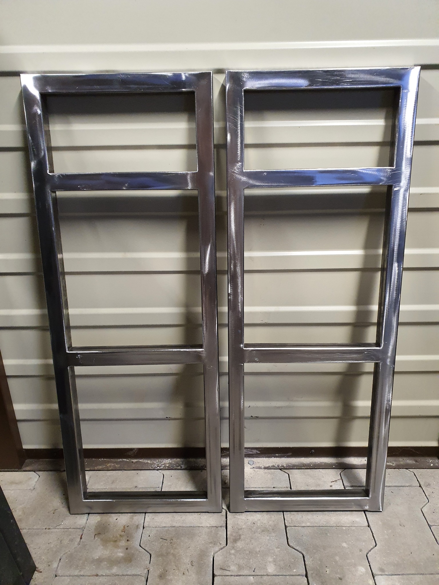 Dwie surowe, metalowe ramy z profili kwadratowych, prawdopodobnie do balustrady lub ogrodzenia, widoczne ślady spawania i obróbki, tło stanowi blaszana ściana garażu.