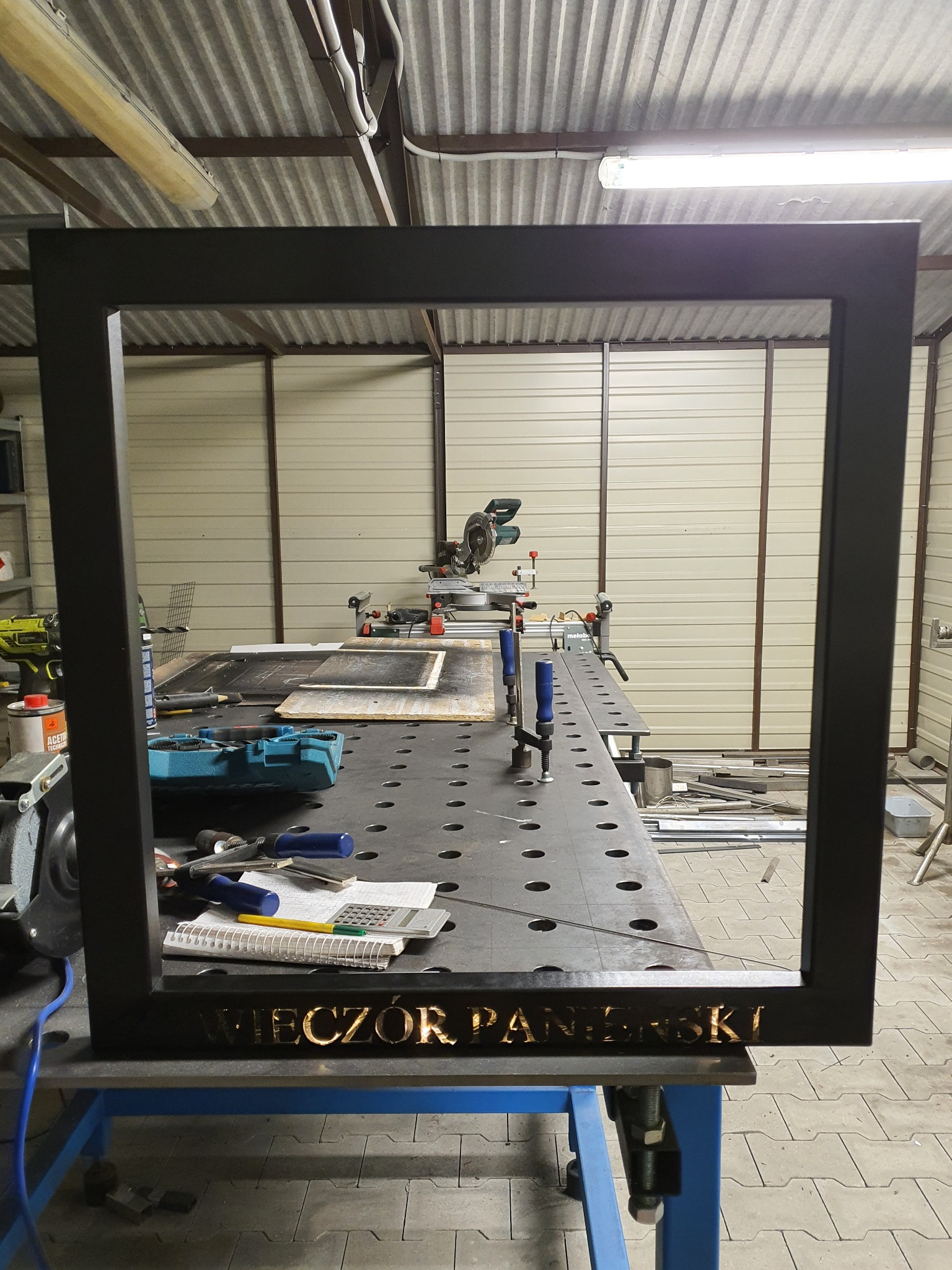Czarna, metalowa rama z wyciętym napisem 'WIECZÓR PANIEŃSKI' stoi na stole warsztatowym, w tle narzędzia i piła do metalu.