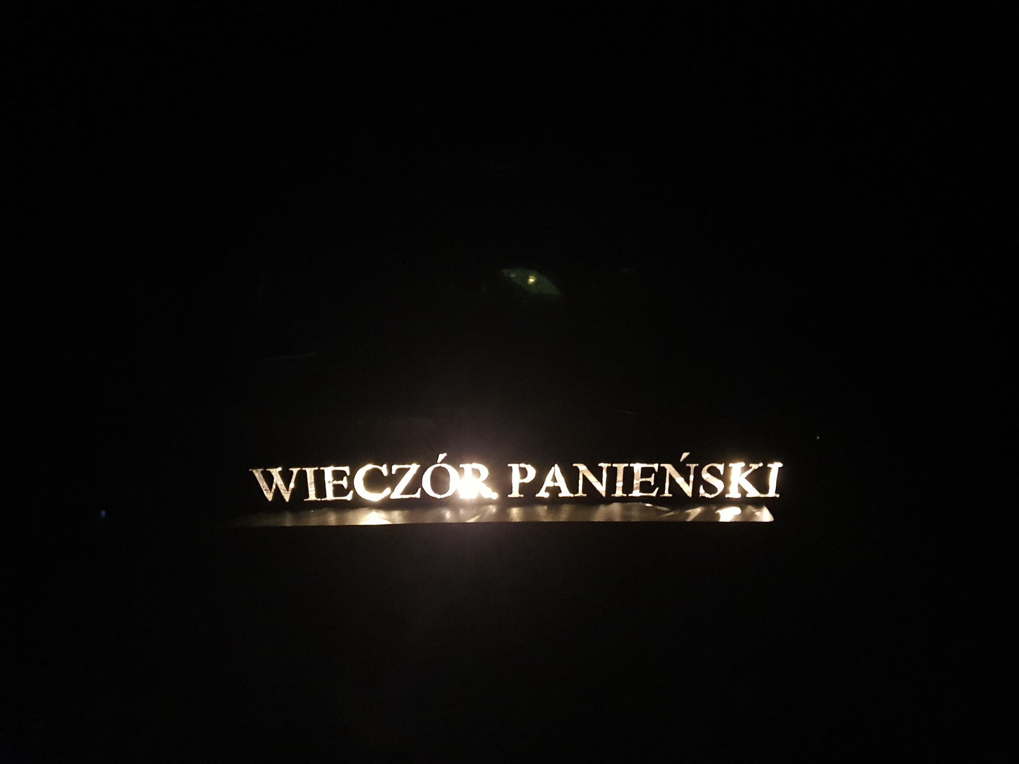 Podświetlany napis 'WIECZÓR PANIEŃSKI' w ciemności, oświetlony od dołu, widoczne odbicia światła.