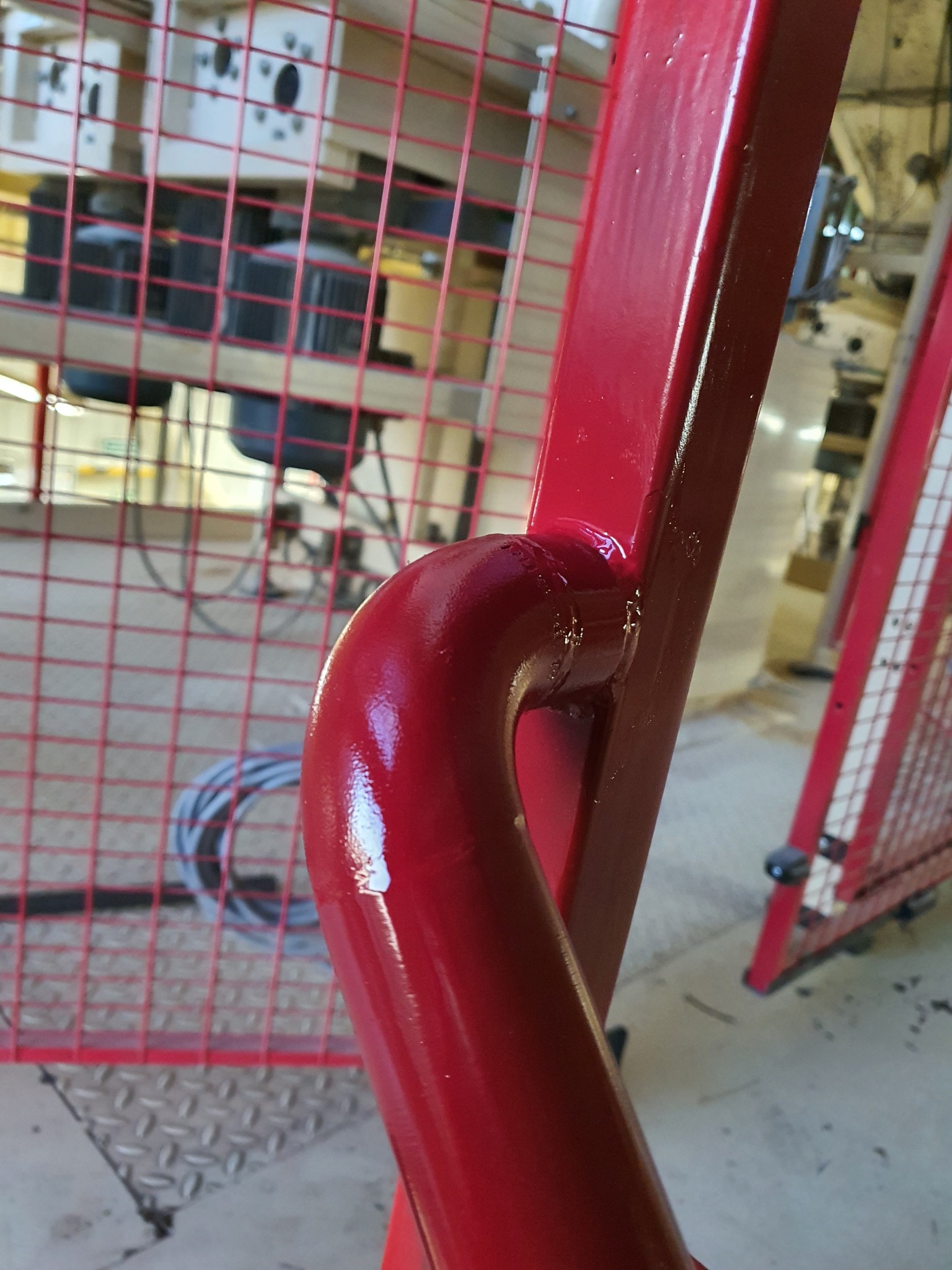 Czerwona, błyszcząca, metalowa konstrukcja ochronna z kratką, prawdopodobnie spawana, z widocznymi śladami spawania i niedoskonałościami farby, na tle podłogi z blachy ryflowanej i fragmentów maszyn.