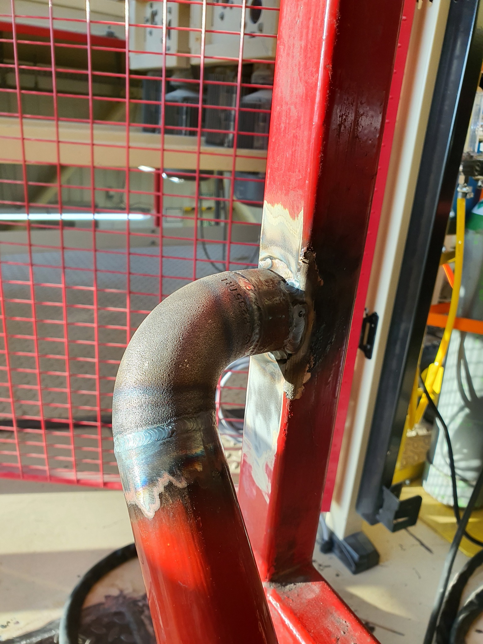 Spoina łącząca metalowy łuk z czerwoną konstrukcją, widoczne ślady po spawaniu, w tle siatka ochronna i elementy warsztatu.