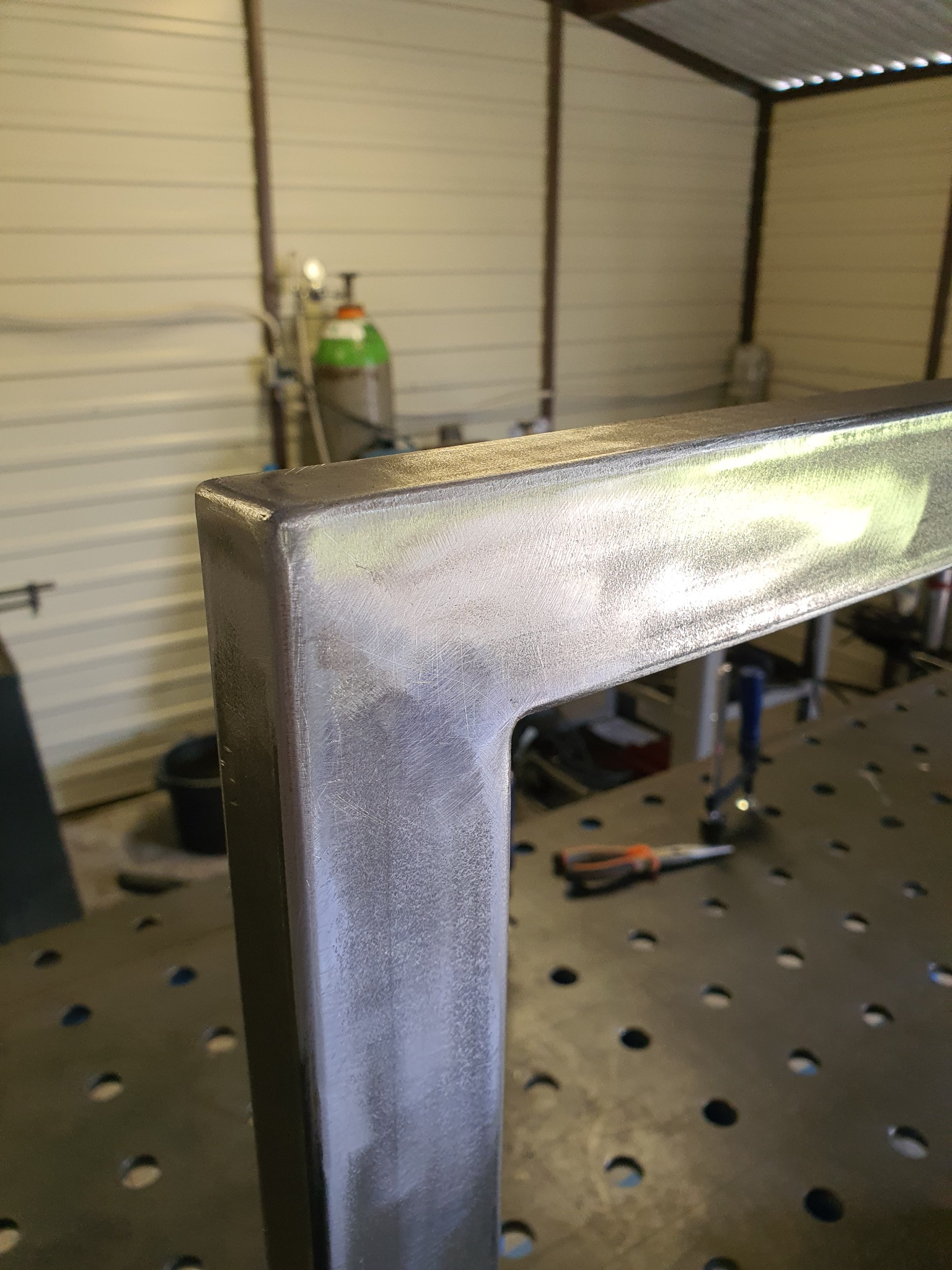 Metalowa rama z profili zamkniętych, świeżo po obróbce, leżąca na perforowanym stole spawalniczym, widoczne narzędzia i butla z gazem w tle.