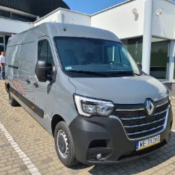 Szary Renault Master, zaparkowany na brukowanym placu przed nowoczesnym budynkiem, z widoczną tablicą rejestracyjną i odblaskami słońca na karoserii.