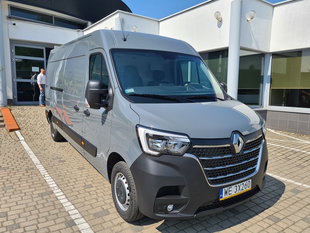 Szary Renault Master, zaparkowany na brukowanym placu przed nowoczesnym budynkiem, z widoczną tablicą rejestracyjną i odblaskami słońca na karoserii.
