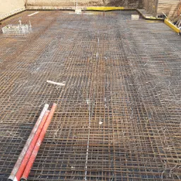 Siatka zbrojeniowa ułożona na czarnej folii, przygotowana do zalania betonem pod płytę fundamentową, widoczne szalunki i wystające rury.