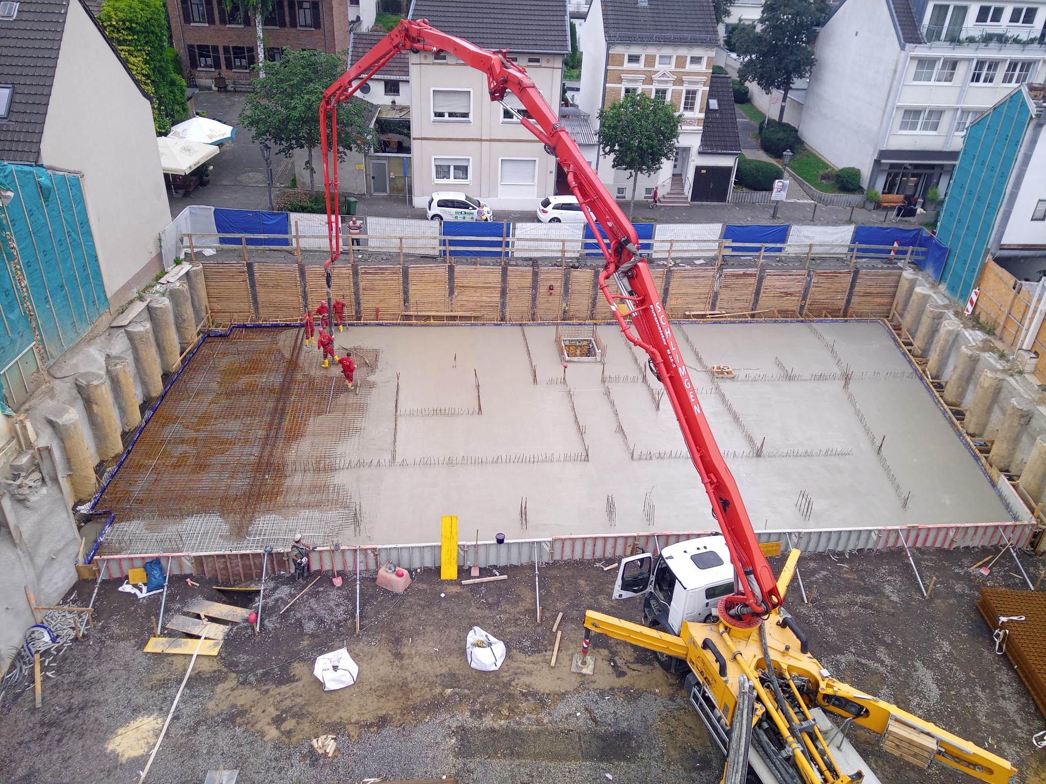 Zalewanie betonem zbrojonej płyty fundamentowej, widok z góry na plac budowy z pompą do betonu i pracownikami w czerwonych kombinezonach. W tle zabudowa miejska.