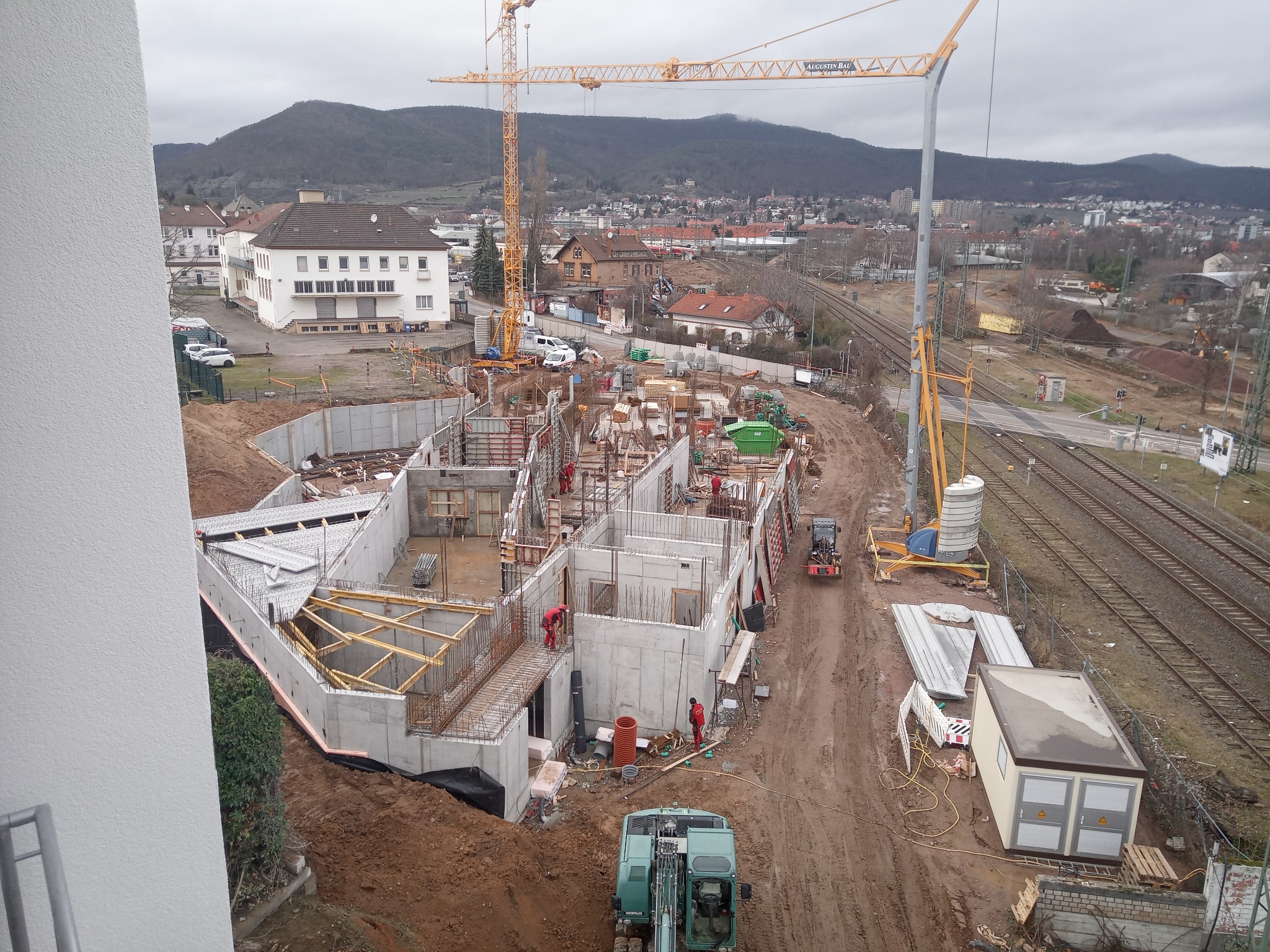 Budowa fundamentów budynku w trakcie realizacji, widoczne szalunki, zbrojenia, pracownicy w czerwonych kombinezonach, dźwig budowlany w tle, teren budowy z koleją obok.