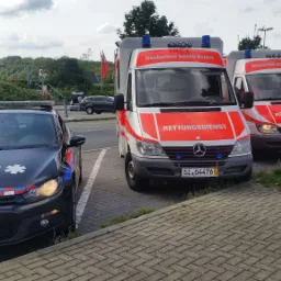 Dwa ambulanse Deutsches Rotes Kreuz i oznakowany samochód policyjny na parkingu, widoczne numery rejestracyjne pojazdów.