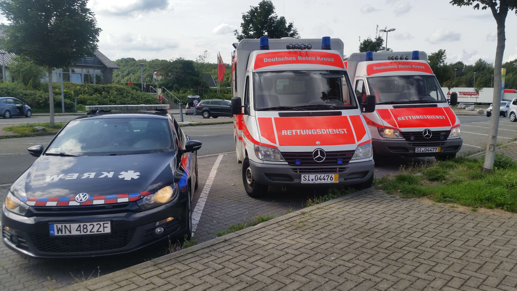 Dwa ambulanse Deutsches Rotes Kreuz i oznakowany samochód policyjny na parkingu, widoczne numery rejestracyjne pojazdów.