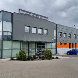 Nowoczesny budynek biurowy z napisem 'bramy drzwi ogrodzenia' na dachu, szaro-pomarańczowa elewacja, parking z samochodami, kostka brukowa, pochmurne niebo.