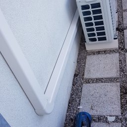 OLINA Sp. z o.o. - Biała jednostka zewnętrzna klimatyzatora zamontowana na ścianie budynku obok rynny, na podłożu z kamiennych płyt i żwiru, obok niebieska pompa próżniowa i papierowa torba.