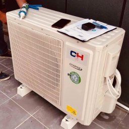 OLINA Sp. z o.o. - Zewnętrzna jednostka klimatyzacji na białych wspornikach, z silikonem, telefonem i dokumentami na górze, po instalacji na szarej podłodze.