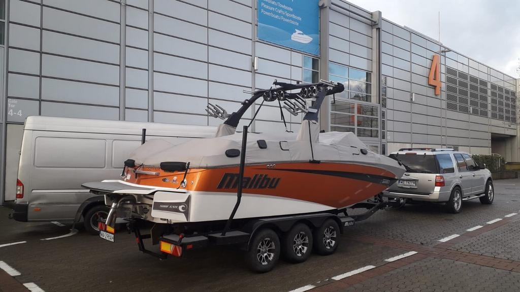 Pomarańczowo-biała łódź Malibu na przyczepie holowana przez srebrny SUV na tle nowoczesnego budynku z numerem 4 i zaparkowanym białym busem.