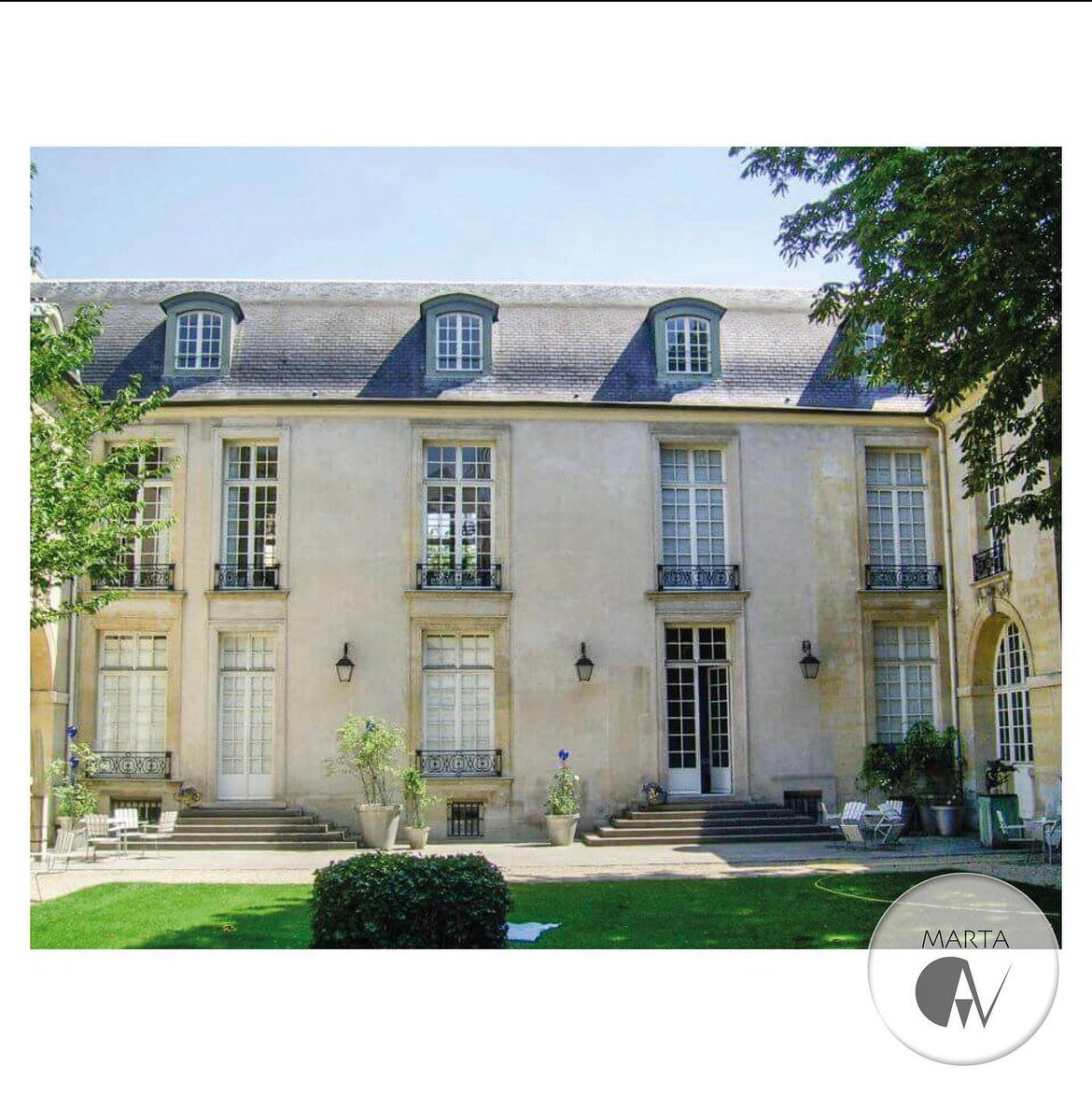 Elewacja budynku usługowego w stylu klasycystycznym z symetrycznym układem okien, mansardowym dachem i zielonym trawnikiem przed wejściem.