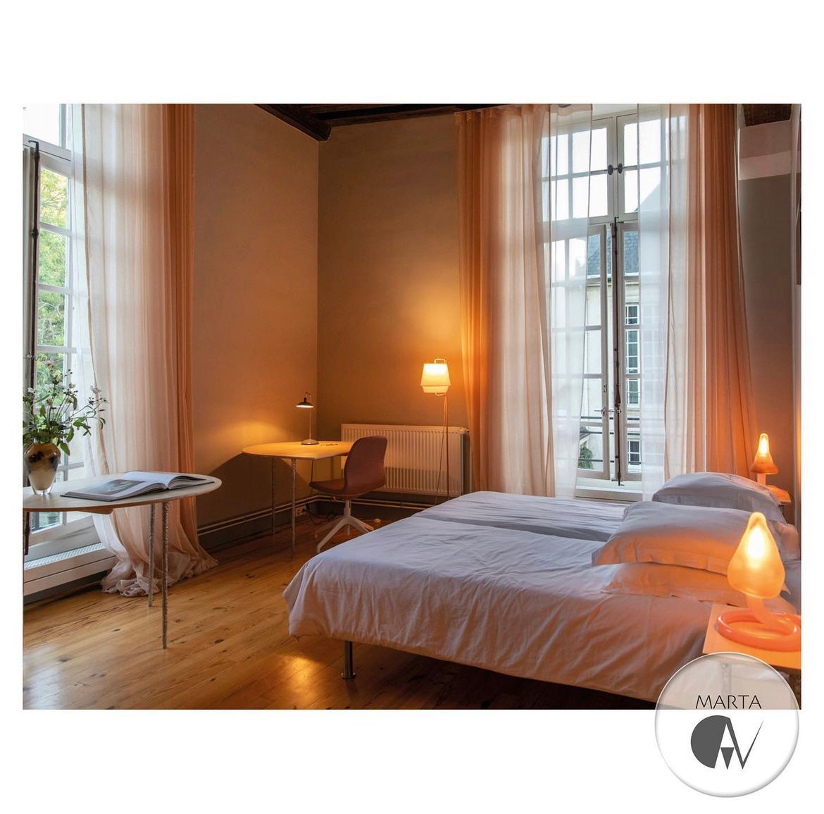 Jasna sypialnia z dwoma pojedynczymi łóżkami, biurkiem przy oknie i kwiatami w wazonie. Ciepłe oświetlenie z lampki nocnej i lampy podłogowej. Widoczne drewniane podłogi i kremowe zasłony.