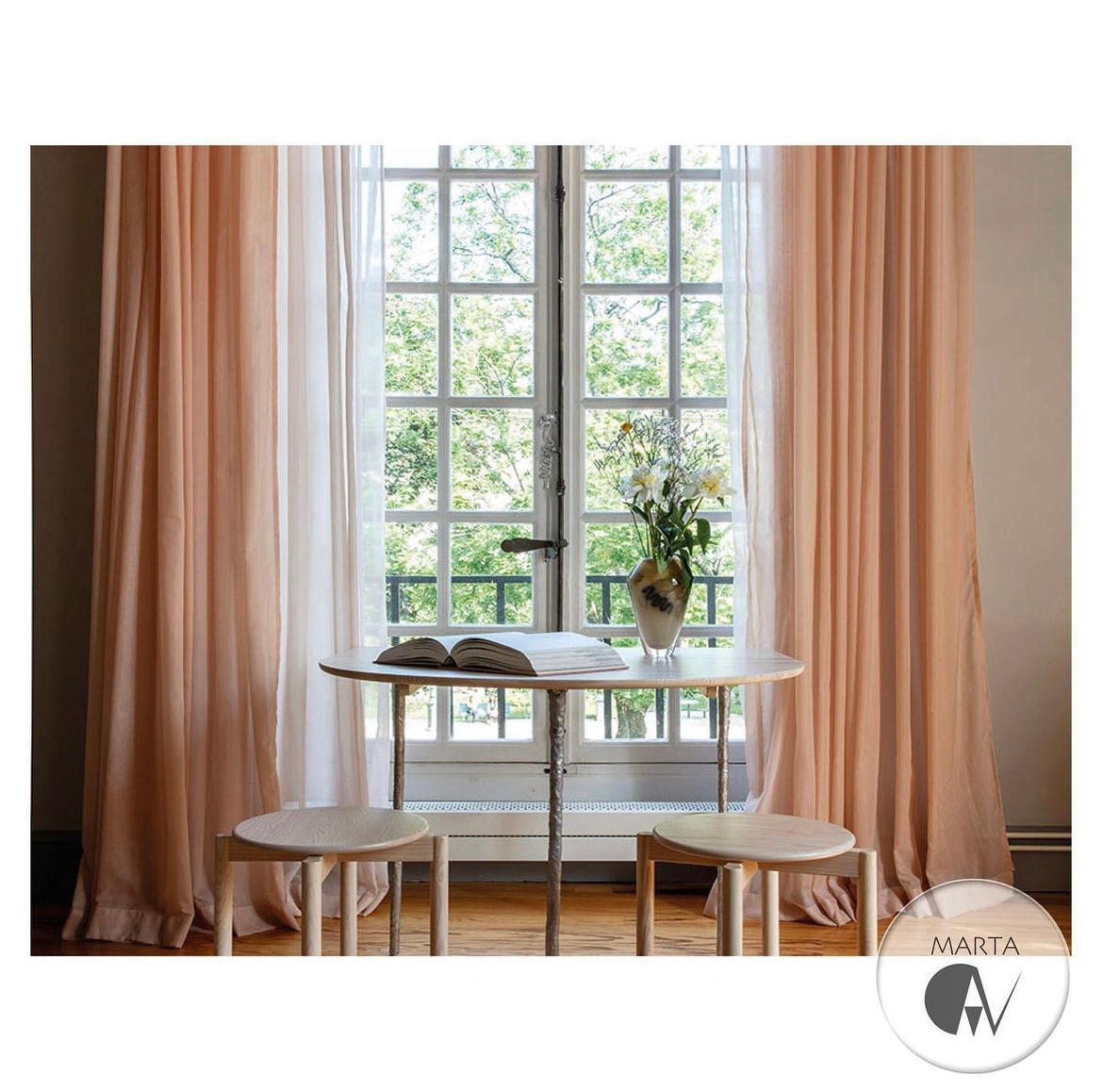 Jasny pokój z oknem: okrągły stół z otwartą książką i wazonem kwiatów, dwa drewniane stołki, zwiewne brzoskwiniowe zasłony, widok na zieleń za oknem.