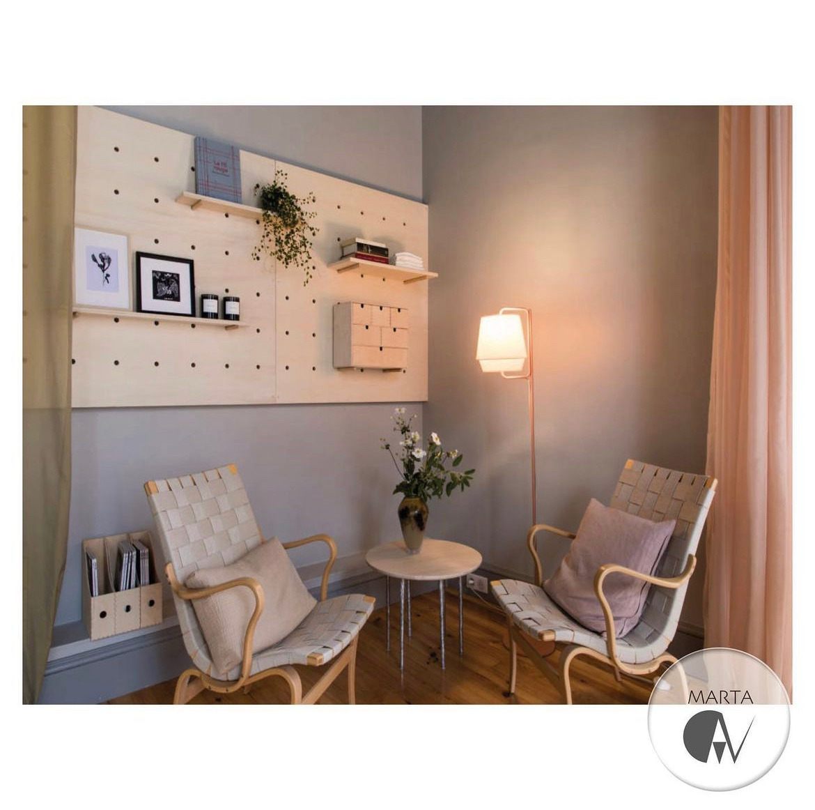 Ustawienie dwóch foteli z plecionymi siedziskami i drewnianymi ramami wokół okrągłego stolika z kwiatami, podświetlonych lampą podłogową, na tle ściany z drewnianymi półkami i dekoracjami.