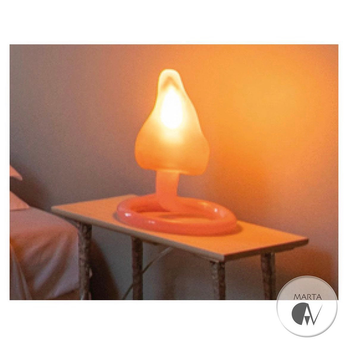 Nietypowa lampka nocna w kształcie grzyba z różową podstawą, stojąca na stoliku nocnym obok łóżka; ciepłe, rozproszone światło.