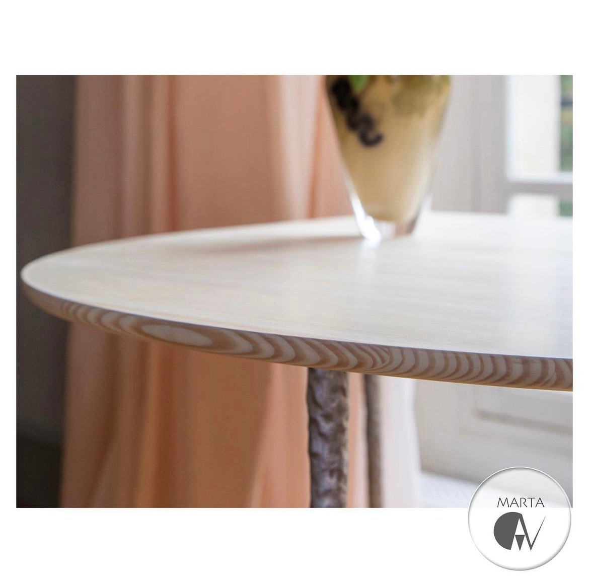 Blat okrągłego stolika z jasnego drewna z widocznym usłojeniem, dekoracyjny wazon z deserem w tle, logo firmy w prawym dolnym rogu.