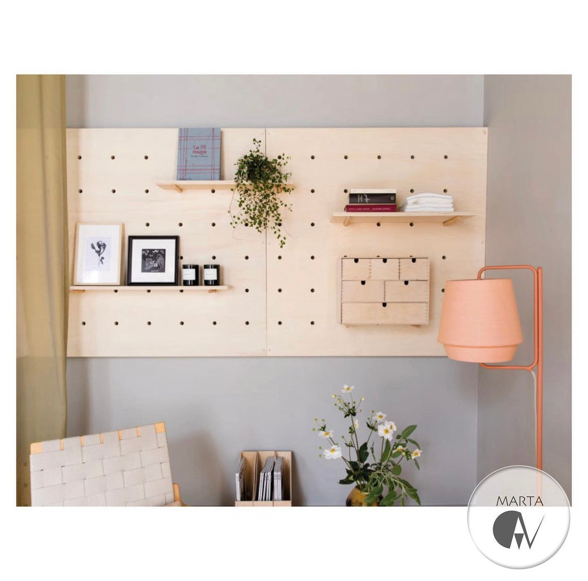 Płyta perforowana z jasnego drewna na ścianie, z półkami, książkami, rośliną doniczkową, ramkami ze zdjęciami i małą komodą. Obok stoi lampa podłogowa z abażurem w odcieniach brzoskwini, a w tle...