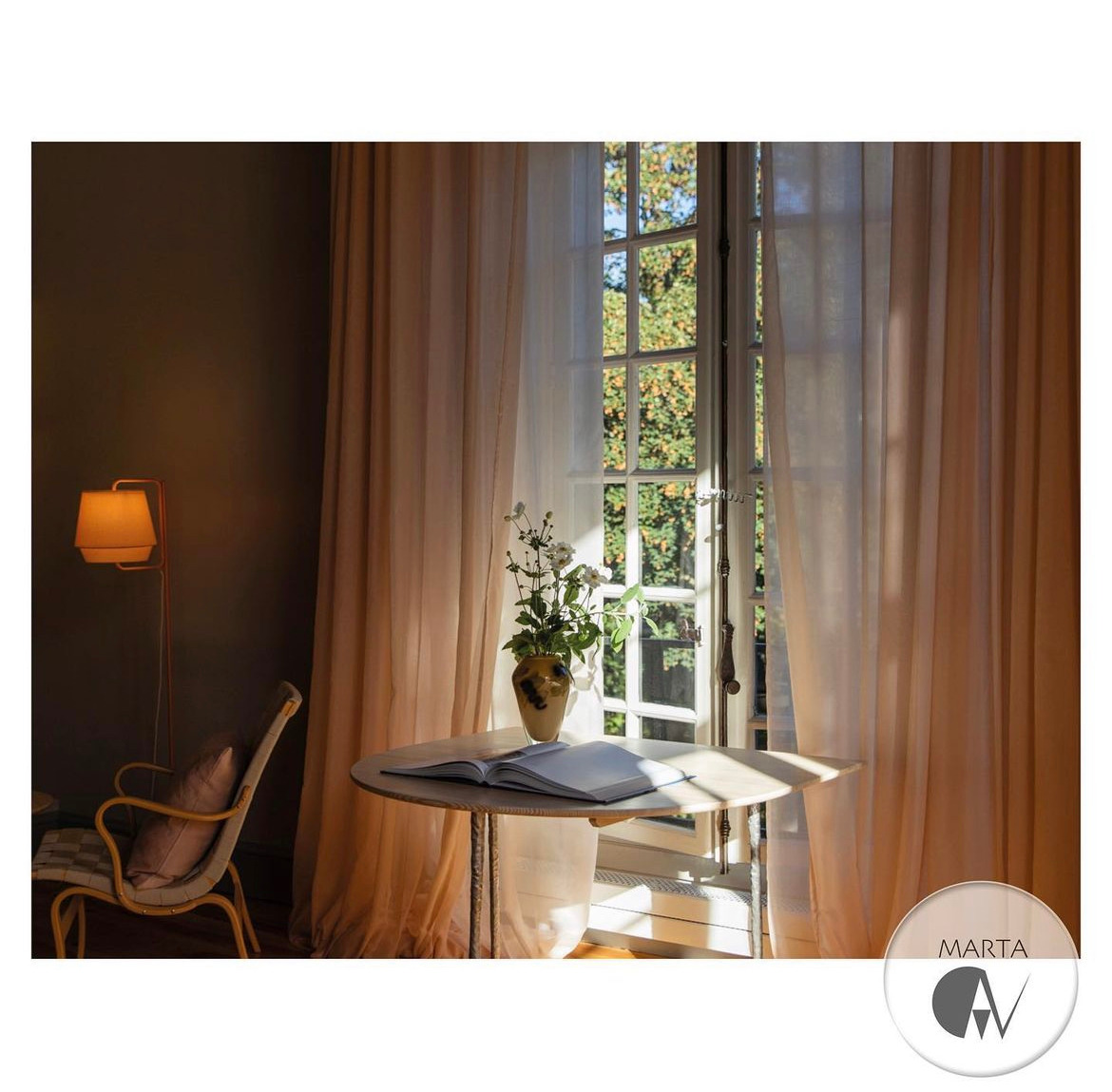 Jasny pokój z okrągłym stolikiem, wazonem z kwiatami i otwartą książką, w tle okno z widokiem na zieleń i zwiewne, beżowe zasłony, obok lampa podłogowa i fotel z poduszką.