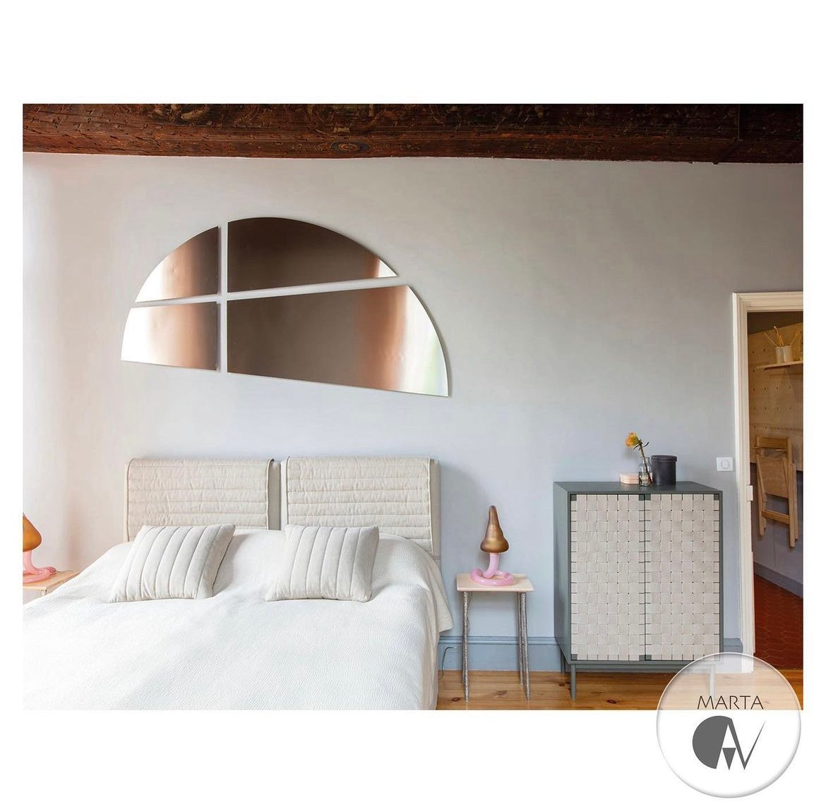 Sypialnia w jasnych odcieniach szarości z łóżkiem z pikowanym zagłówkiem, nad którym wisi dekoracyjne lustro w kształcie półkola podzielonego na cztery segmenty, obok szafka z plecionymi drzwiami...