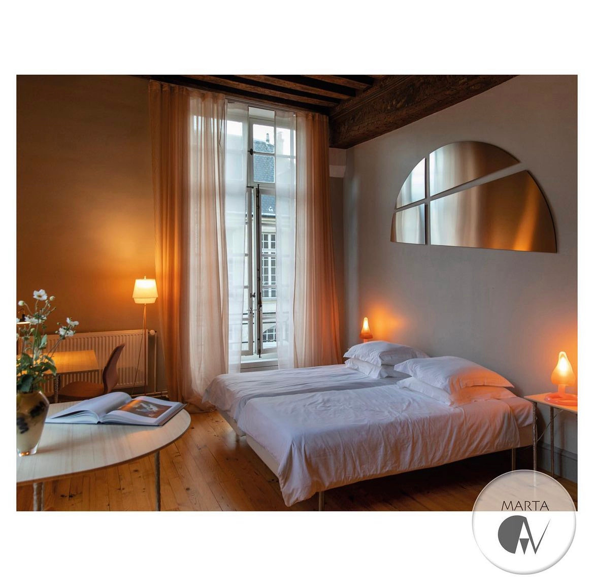 Pokój hotelowy z dwoma pojedynczymi łóżkami, drewnianą podłogą, oknem z widokiem na budynki i dekoracją ścienną w kształcie półksiężyca. Na stoliku nocnym lampka w kolorze pomarańczowym.