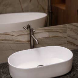 MD CONCEPT - Owalna, biała umywalka nablatowa na ciemnym blacie z widoczną baterią i fragmentem wanny w tle, w łazience wykończonej marmurem.