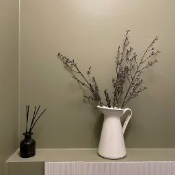 Minimalistyczna łazienka: biały dzbanek z gałązkami i czarny dyfuzor zapachowy na tle oliwkowej ściany i białych, wąskich płytek.