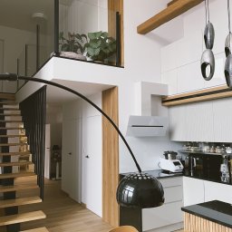 MD CONCEPT - Dwupoziomowe mieszkanie: jasna kuchnia z drewnianymi akcentami, schody z czarną balustradą i antresola z roślinami. Nowoczesny wystrój.