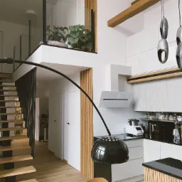Dwupoziomowe mieszkanie: jasna kuchnia z drewnianymi akcentami, schody z czarną balustradą i antresola z roślinami. Nowoczesny wystr&oacute;j.