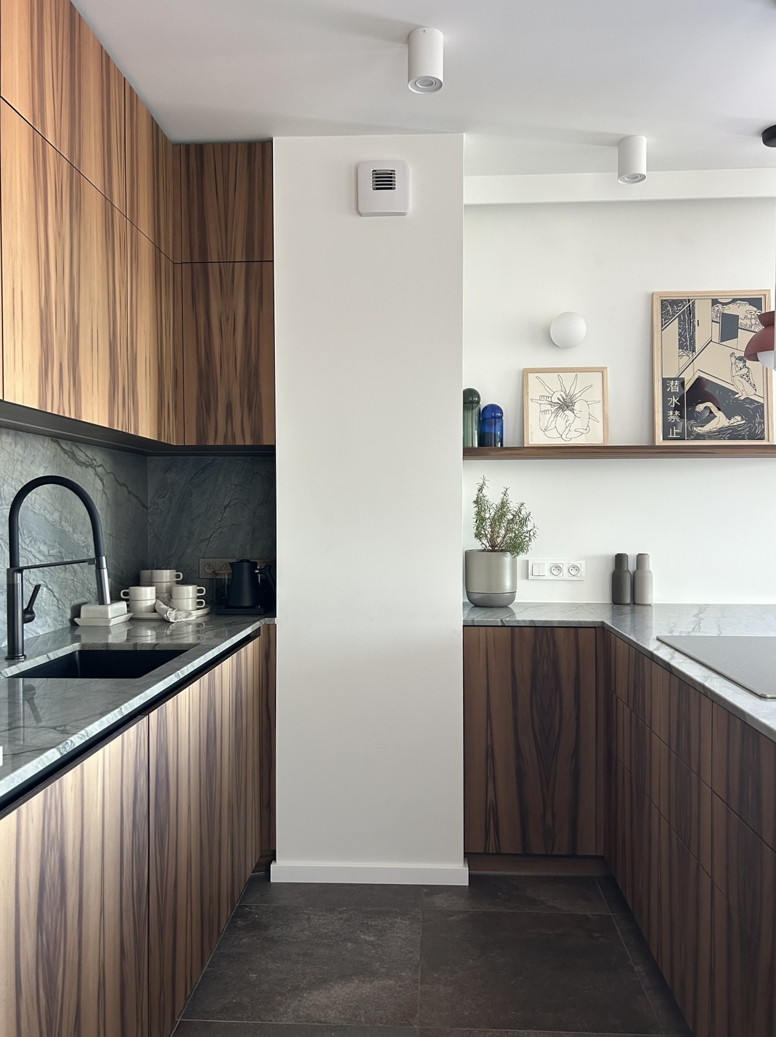 Nowoczesna kuchnia z drewnianymi frontami, marmurowym blatem i czarną armaturą. Minimalistyczny wystrój z półką i grafikami na ścianie.