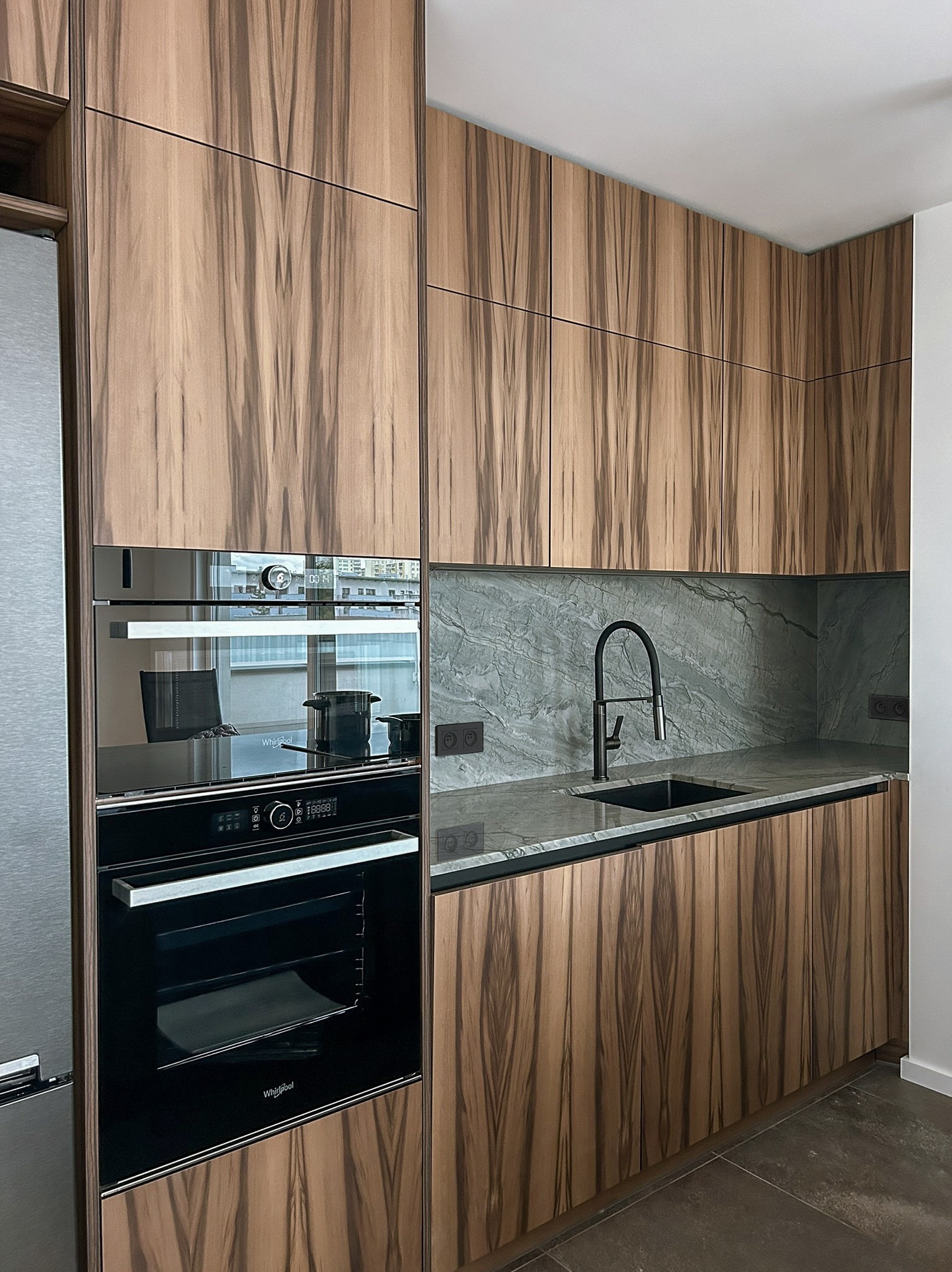 Nowoczesna kuchnia z fornirem w zebrę: zabudowa z piekarnikiem Whirlpool, czarna armatura i blat z szarego marmuru. Minimalistyczny design i eleganckie wykończenie.