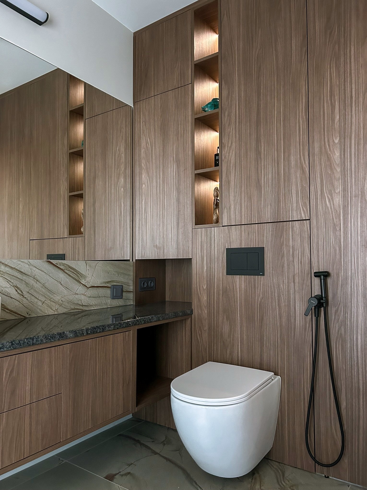 Nowoczesna łazienka z drewnianą zabudową, podświetlanymi półkami i marmurowym blatem umywalki. Minimalistyczny design, wisząca toaleta, bidetka.