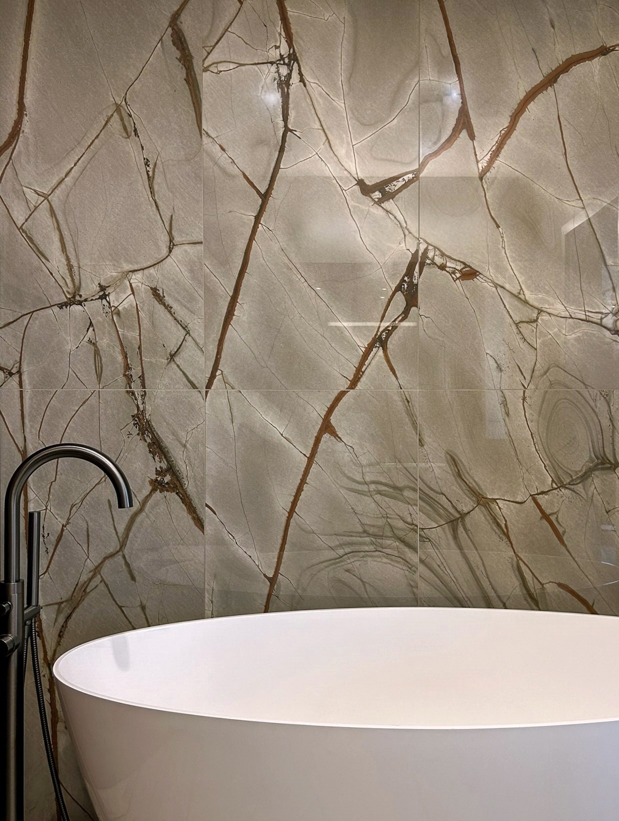 Biała wanna wolnostojąca z chromowaną baterią na tle ściany wyłożonej płytkami imitującymi marmur z wyraźnym, brązowym żyłowaniem.