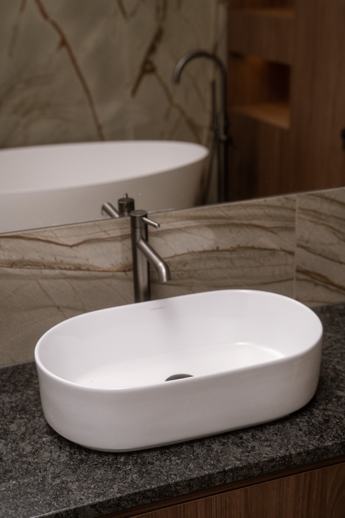 Owalna, biała umywalka nablatowa na ciemnym blacie z widoczną baterią i fragmentem wanny w tle, w łazience wykończonej marmurem.