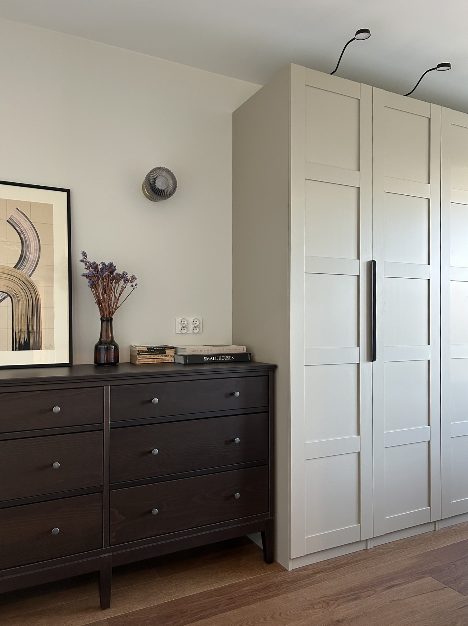 Sypialnia w neutralnych barwach: ciemna komoda z dekoracjami, jasna szafa z długimi uchwytami i minimalistyczne oświetlenie sufitowe.